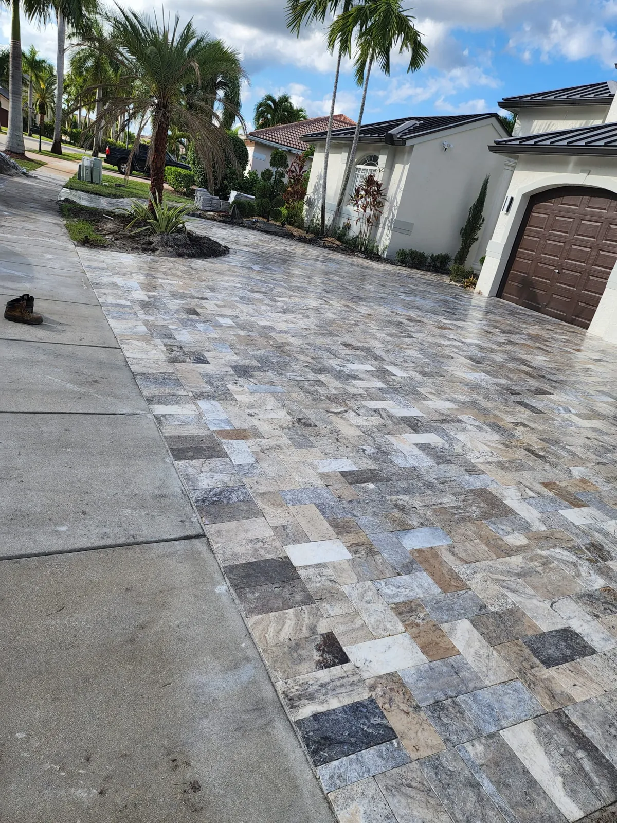 Paver driveway pattern meeting a concrete strip, with palms and a home behind