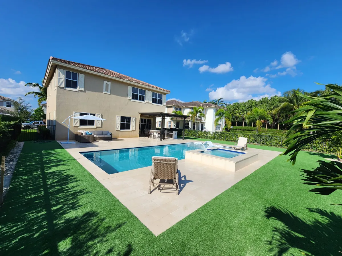Pale stone terrace around a pool and raised spa, with a turf lawn and a two-story home behind
