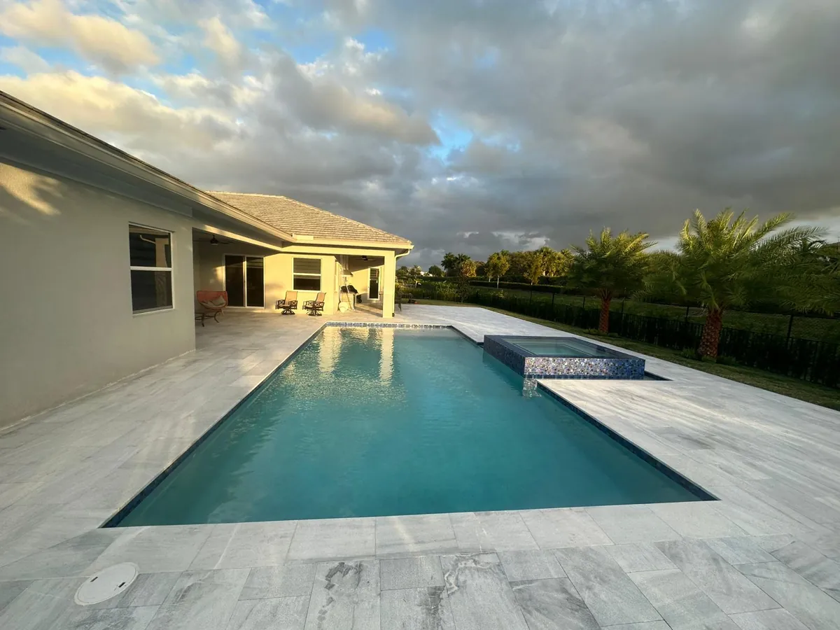 Pool with a pale stone deck and covered patio under an evening sky