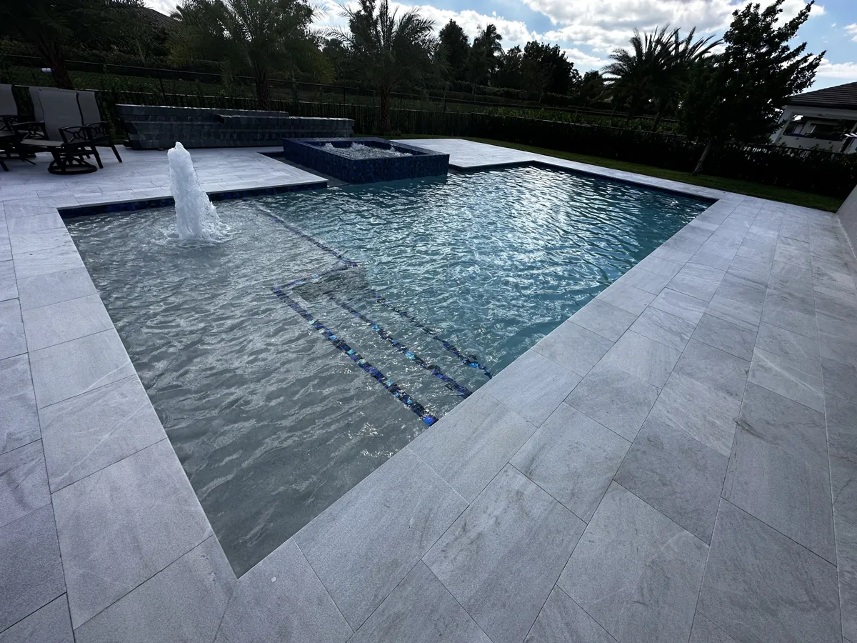 Rectangular pool with blue tile-lined steps and a water jet, surrounded by gray paving