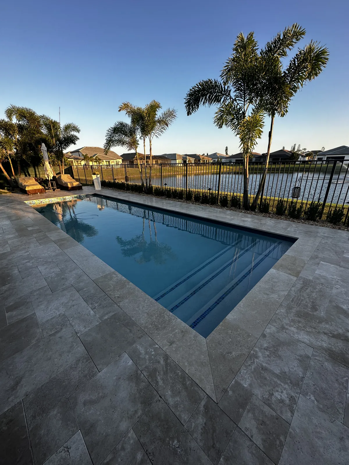 Gray stone pool deck at sunset beside an iron fence, palms and open water