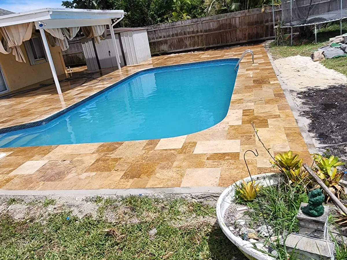 Warm travertine-toned paving around a blue pool in a fenced backyard
